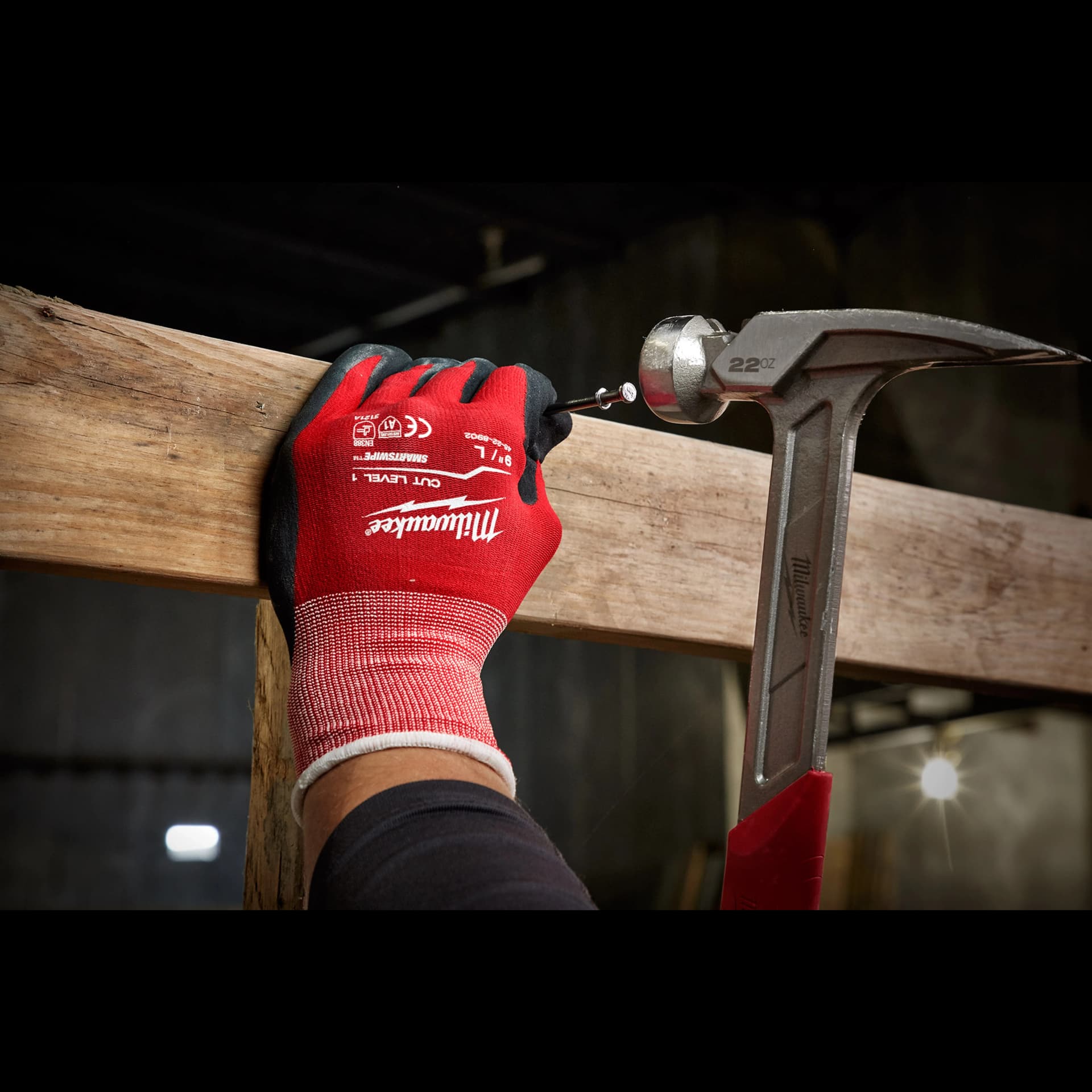 A person wearing red and black Cut%201%20Dipped%20Gloves is holding a wooden plank while using a hammer to drive a screw into it. The scene appears to be in a dimly lit, industrial environment.