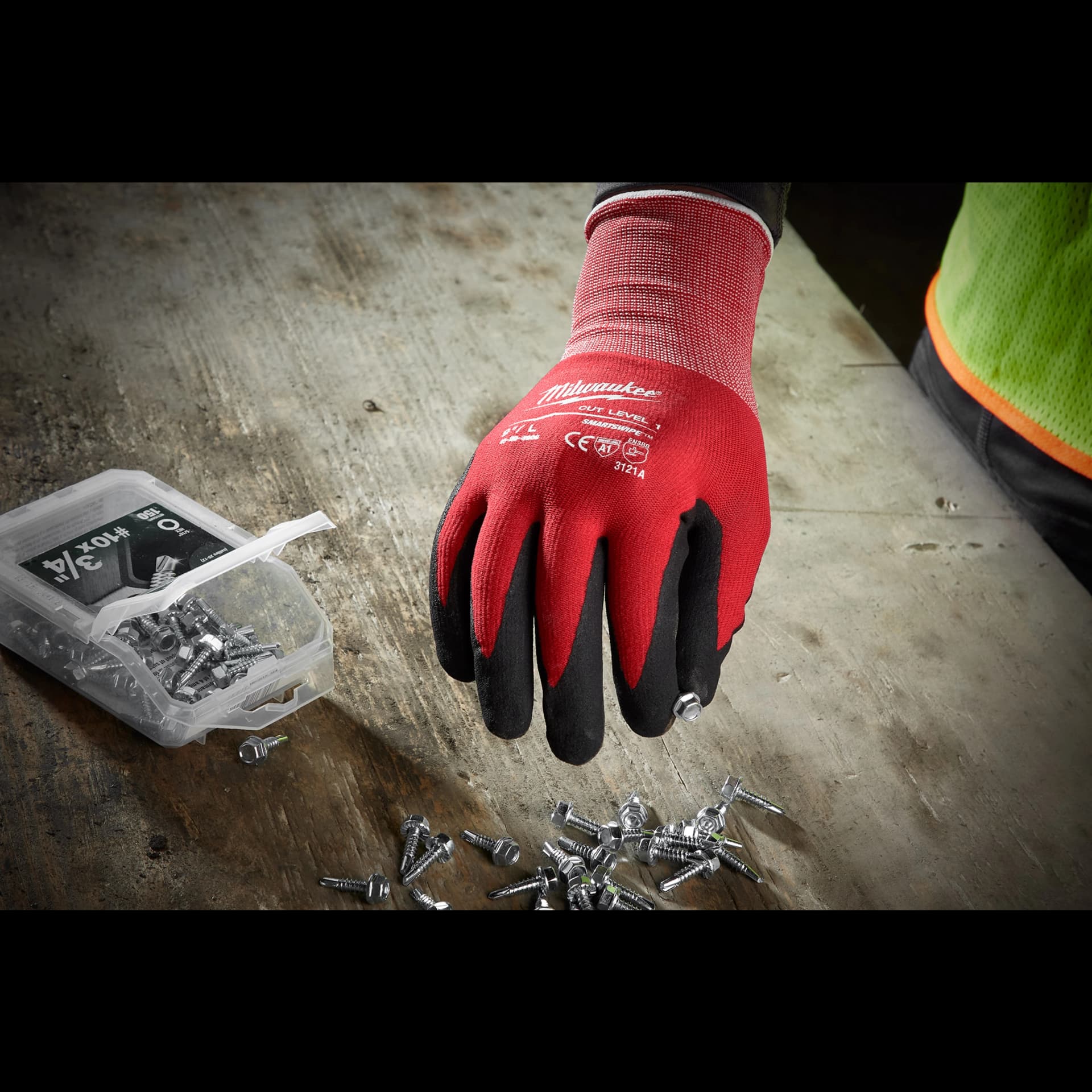 A person wearing Milwaukee Cut 1 Dipped Gloves is picking up screws from a wooden surface next to an open plastic container filled with screws. The gloves are red with black fingertips and have protective labels on the back.